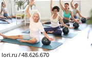 Concentrated young adult Hispanic woman, fitness enthusiast, participating group Pilates session in cozy sunlit studio, doing side bend with exercise ball, promoting balance and lateral stretching. Стоковое видео, видеограф Яков Филимонов / Фотобанк Лори