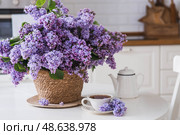 A large purple bouquet of lilacs in a wicker basket, a teapot and a cup of tea on the table. In the background is the interior of a modern white kitchen. The concept of home comfort. Стоковое фото, фотограф Photojuli / Фотобанк Лори