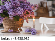 A large purple bouquet of lilacs in a wicker basket, a cup of tea and candles on the table. In the background is the interior of a modern white kitchen. The concept of home comfort. Стоковое фото, фотограф Photojuli / Фотобанк Лори