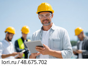 A man wearing a yellow hard hat is holding a tablet in front of a group of const. Стоковое AI-изображение, AI-художник Olga Golub / Фотобанк Лори