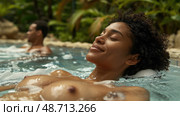A woman enjoys a peaceful soak in warm water surrounded by lush foliage, while another person relaxes nearby, soaking in the ambiance. Стоковое AI-изображение, AI-художник Данила Большаков / Фотобанк Лори