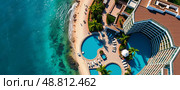 Aerial view of a coastal resort with turquoise swimming pools, palm trees, sandy beach, and ocean waves. Guests relax on sun loungers under umbrellas, creating a tropical paradisiacal scene. Стоковое AI-изображение, AI-художник Olga Dmitrieva / Фотобанк Лори