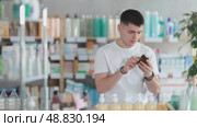 Guy in a summer t-shirt is standing near the pill racks and choosing a medicinal cough syrup at the pharmacy. Young man buying medicine for cold and flu symptoms. Стоковое видео, видеограф Яков Филимонов / Фотобанк Лори