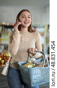Positive young woman looking for something, holding cart with food products and talking on mobile phone at store. Стоковое видео, видеограф Яков Филимонов / Фотобанк Лори