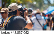 A photographer in a tan cap focuses on taking a photograph at a public event, surrounded by a crowd of people. Стоковое AI-изображение, AI-художник Данила Большаков / Фотобанк Лори