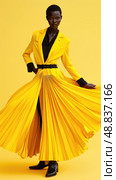 A woman in a bright yellow pleated dress poses against a vibrant yellow backdrop. The dress has a cinched waist with a black belt and the skirt is dramatically flared. Стоковое AI-изображение, AI-художник Данила Большаков / Фотобанк Лори
