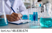 Laboratory technician conducting experiments with blue liquid in glassware, focused on recording observations. Стоковое AI-изображение, AI-художник Сергей Толмачёв / Фотобанк Лори