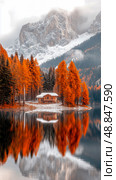 Autumn reflection: A quaint cabin beside a still lake mirroring vibrant fall foliage against a backdrop of snow-dusted mountains. Стоковое AI-изображение, AI-художник Данила Большаков / Фотобанк Лори