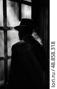 A man in a hat stands by a window, his face obscured by shadow, looking out. He seems contemplative, lost in thought. Стоковое AI-изображение, AI-художник Данила Большаков / Фотобанк Лори