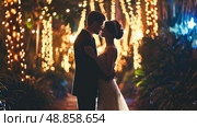 A bride and groom stand close together, embracing under a canopy of twinkling string lights in a garden setting at night. Стоковое AI-изображение, AI-художник Данила Большаков / Фотобанк Лори