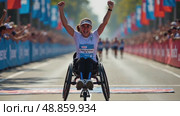 A woman in a wheelchair raises her right arm in a fist pump as she crosses the finish line of a marathon. The background is filled with spectators cheering the winner. Parasports competitions. Стоковое AI-видео, аниматор Sergey Grishin / Фотобанк Лори
