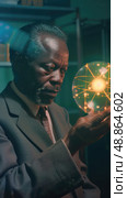A senior scientist with a graying beard studies an atom model while sitting in a dimly lit laboratory. Стоковое AI-изображение, AI-художник Данила Большаков / Фотобанк Лори