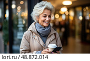 Senior woman effortlessly balances a steaming cup of coffee in one hand, while skillfully navigating her cell phone with the other. Стоковое AI-изображение, AI-художник Данила Большаков / Фотобанк Лори