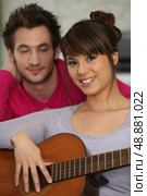 smiling brunette playing the guitar under boyfriend&rsquo;s watchful eye. Стоковое фото, агентство Ingram Publishing / Фотобанк Лори