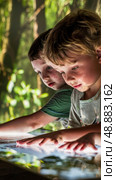 Two young boys are engrossed in an interactive exhibit in a forest setting. They are touching the screen with fascination and exploring the world around them. Стоковое AI-изображение, AI-художник Данила Большаков / Фотобанк Лори