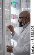 A pharmacist in a white coat retrieves medication from shelves in a pharmacy. Стоковое AI-изображение, AI-художник Данила Большаков / Фотобанк Лори