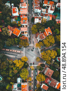 An aerial view of a city intersection with cars driving on the roads and buildings on either side. The trees are in full bloom and the city looks bustling. Стоковое AI-изображение, AI-художник Данила Большаков / Фотобанк Лори