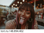 A close-up photo of a woman with long, dark hair and pink highlights, wearing a sequined top, and sticking her tongue out playfully as she smiles in front of a blurry background of lights at night. Стоковое AI-изображение, AI-художник Данила Большаков / Фотобанк Лори