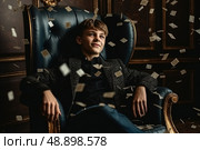 Teen boy is depicted sitting in a chair, surrounded by piles of money, symbolizing his immersion in wealth and prosperity. Стоковое AI-изображение, AI-художник Данила Большаков / Фотобанк Лори