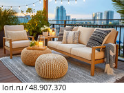 Cozy outdoor lounge area featuring stylish seating with cushions, decorative poufs, and ambient lighting, surrounded by greenery and modern architecture, perfect for relaxation and social gatherings. Стоковое AI-изображение, AI-художник Ирина Ручушкина / Фотобанк Лори