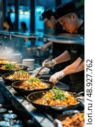 A chef in a black uniform serves stir-fried dishes from a restaurant buffet. The food is arranged in a row of black woks and appears to be steaming. Стоковое AI-изображение, AI-художник Данила Большаков / Фотобанк Лори