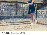 A young woman wearing a blue dress is standing on the beach on a sunny day by an old wooden structure with rusty chains hanging from it. Стоковое фото, агентство Ingram Publishing / Фотобанк Лори