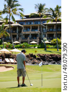 A man prepares to putt on a golf course with a luxury resort in the background. He is wearing khaki shorts and a green striped shirt. Palm trees and lush green foliage are visible. Стоковое AI-изображение, AI-художник Данила Большаков / Фотобанк Лори