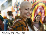 A young woman with a shaved head smiles brightly, wearing a plaid shirt and backpack. She is standing with a group of friends in an urban setting with colorful murals in the background. Стоковое AI-изображение, AI-художник Данила Большаков / Фотобанк Лори