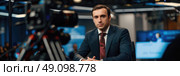 A man in a suit sits in a news studio, looking at the camera with a serious expression. The studio is lit with bright lights, and there are screens behind him displaying news headlines. Стоковое AI-изображение, AI-художник Данила Большаков / Фотобанк Лори