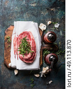 Raw marbled meat steaks with wrapping paper on rustic cutting board with fresh flavoring and spice mills on dark rustic background, top view. Стоковое фото, агентство Ingram Publishing / Фотобанк Лори