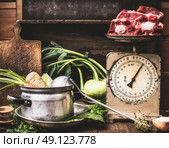 Kitchen table with cooking pot, ladle, vegetables and old weigher with raw meat , preparation of soup , broth or stew, front view.  Country style. Стоковое фото, агентство Ingram Publishing / Фотобанк Лори