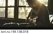 A man sits on a couch, strumming a guitar with focused concentration. Стоковое AI-изображение, AI-художник Данила Большаков / Фотобанк Лори