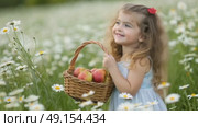 A cheerful girl with long curly hair gathers apples in a woven basket while standing in a beautiful meadow filled with blooming flowers. Стоковое AI-видео, аниматор Оксана Медведева / Фотобанк Лори
