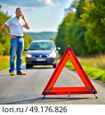 On a calm afternoon, a man stands next to his stalled car on a quiet road, thoughtfully pondering his next move while a bright warning triangle signals drivers to proceed with caution. Стоковое AI-изображение, AI-художник Ольга Игнашина / Фотобанк Лори