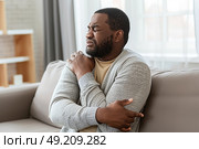 A man sitting on a couch in a living room holds his shoulder in discomfort. He appears to be in pain, possibly from an injury or soreness. Стоковое AI-изображение, AI-художник Данила Большаков / Фотобанк Лори