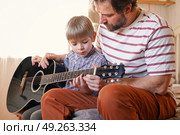 Handsome dad teaches his cute son to play the guitar sitting on sofa in the living room at home. Happy Father's Day. Father teaching his child to play guitar. Kid Learning music skill. Art education. Стоковое фото, фотограф Марина Демидюк / Фотобанк Лори