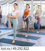 Young women doing Hatha yoga in modern studio, performing balancing asana Vrikshasana with hands clasped in namaste above head. Стоковое фото, фотограф Яков Филимонов / Фотобанк Лори