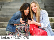 Two young women looking at some funny thing on their smart phone outdoors, sitting on urban steps. Friends girls laughing. Стоковое фото, агентство Ingram Publishing / Фотобанк Лори