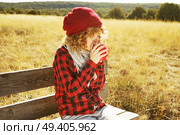 Front portrait of a young woman in red plaid shirt with a wool cap and scarf taking a cup of tea or coffee while she is sunbathing sitting in a wooden bench in a yellow field with backlight from autumn sun. Стоковое фото, агентство Ingram Publishing / Фотобанк Лори