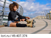 Young smiling black girl sitting on urban bridge and puts on skates. Woman with afro hairstyle rollerblading on sunny day. Стоковое фото, агентство Ingram Publishing / Фотобанк Лори