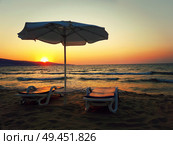 Romantic scene as a sunset at the sea shore with two sunbeds and an umbrella on the sand beach in front of the sun rays. Стоковое фото, агентство Ingram Publishing / Фотобанк Лори