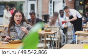 Young woman in a jacket sits on a cool terrace of a restaurant and eats a salad against the background of other visitors. European woman tries a freshly prepared vegetable salad from a chef. Стоковое видео, видеограф Яков Филимонов / Фотобанк Лори