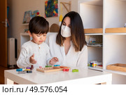 Liltle Preschooler Kid playing tetris wood puzzle with teacher educador help using face mask for coronavirus pandemic. Homeshooling. Learning Community. Montessori School. Стоковое фото, агентство Ingram Publishing / Фотобанк Лори