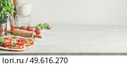 Homemade pizza background with wooden rolling pin, flour and basil leaves on grey kitchen table at white wall background. Cooking Italian food at home. Front view with copy space. Стоковое фото, агентство Ingram Publishing / Фотобанк Лори