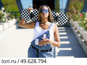 Confident African American female hipster in sunglasses and overalls standing with skateboard in street in summer and looking at camera. Cool black woman with skateboard in city. Стоковое фото, агентство Ingram Publishing / Фотобанк Лори