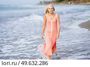 Elderly slim female in maxi summer dress walking in sea water along shore and looking at camera. Senior woman in dress walking along beach. Стоковое фото, агентство Ingram Publishing / Фотобанк Лори