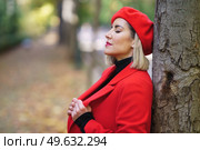 Side view of attractive female in stylish red coat with classic beret leaning on tree trunk with closed eyes in park on blurred background. Dreamy woman near tree in a park. Стоковое фото, агентство Ingram Publishing / Фотобанк Лори