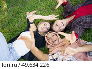 From above of multiracial female friends gesticulating and looking at camera while lying on grassy lawn on summer day in park. Happy diverse women lying on lawn. Стоковое фото, агентство Ingram Publishing / Фотобанк Лори
