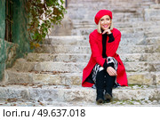 Full body of confident young lady, with blond hair and red lips in elegant coat and beret, sitting on shabby stone stairs with hand at chin and smiling in town. Feminine stylish woman resting on steps in town and smiling. Стоковое фото, агентство Ingram Publishing / Фотобанк Лори