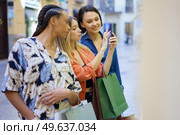 Group of multiethnic female friends taking picture of showcase on smartphone while standing together near boutique on street during shopping. Multiracial women taking photo of showcase in city. Стоковое фото, агентство Ingram Publishing / Фотобанк Лори